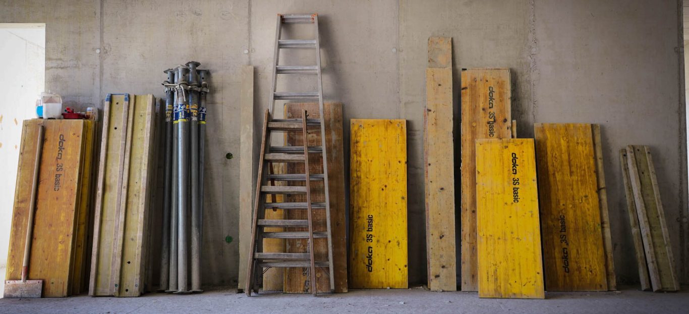 Werkzeuge und Materialien: Holzplatten, Leiter, Rohre, an einer Betonwand gestapelt.
