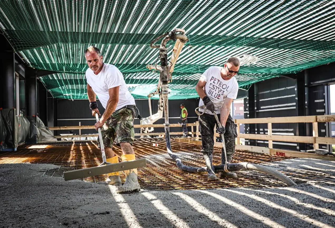 Zwei Bauarbeiter verlegen Beton auf einer Baustelle unter einem grünen Dach.