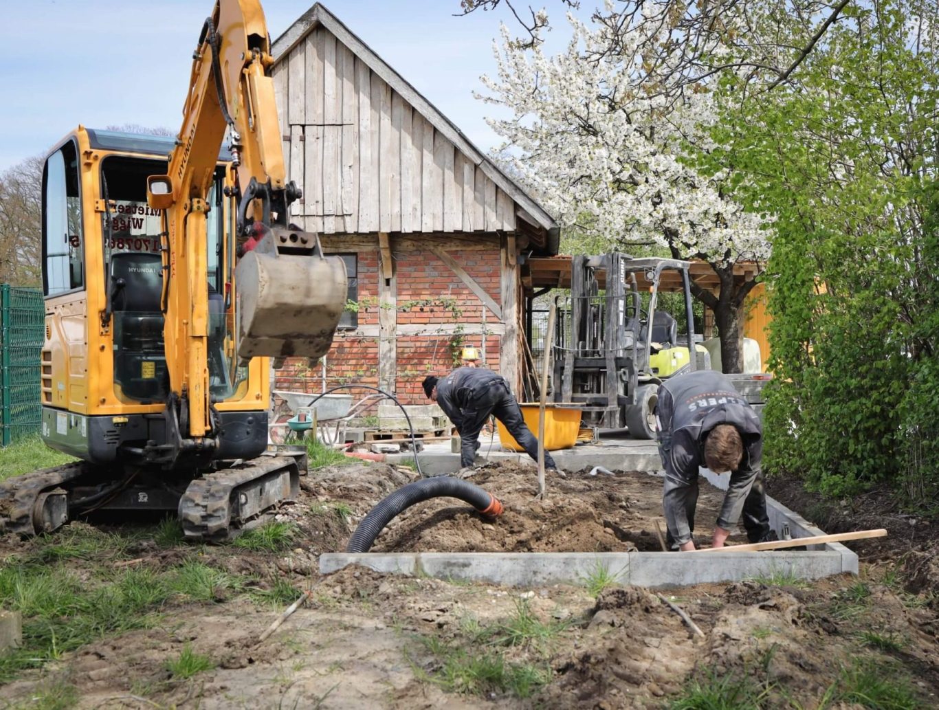 Bauarbeiten mit Bagger und Arbeitern in einem Garten neben einem Haus und blühenden Bäumen.