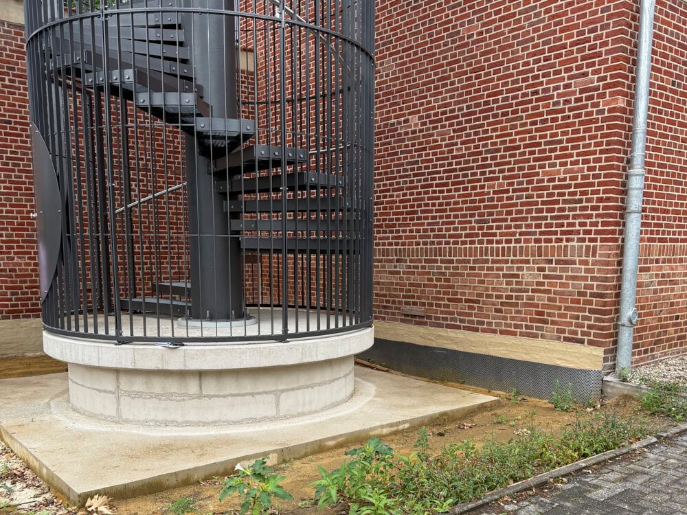 Einfahrt zu einer schwarzen, spiralförmigen Treppe neben einer roten Backsteinmauer.