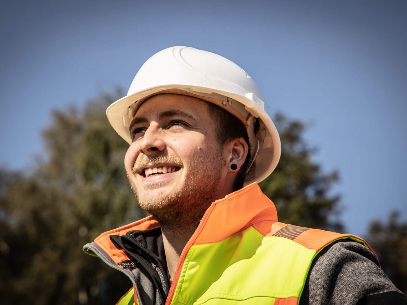 Junger Bauarbeiter in Sicherheitskleidung und Helm, lächelt bei Tageslicht.