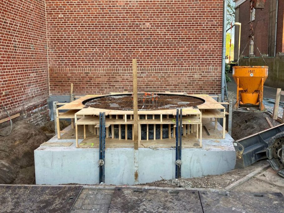 Betonfundament mit Holzrahmen für einen zirkulären Bau, umgeben von Baugeräten.