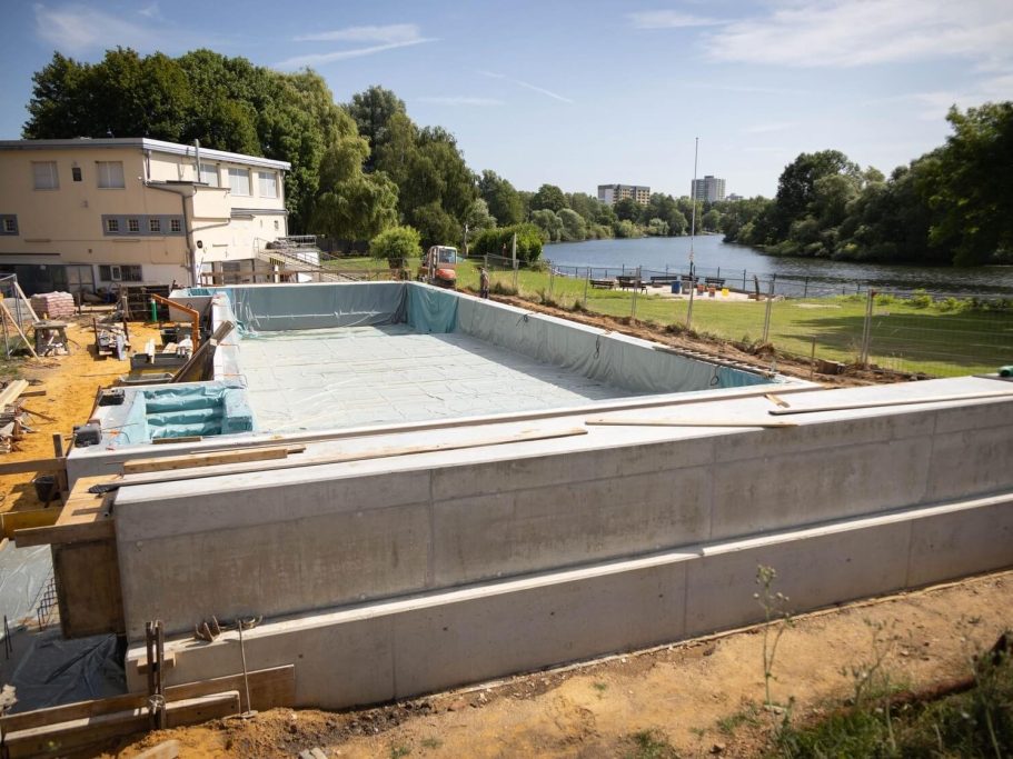 Blick auf einen Pool im Bau neben einem Gewässer und einem angrenzenden Gebäudekomplex.